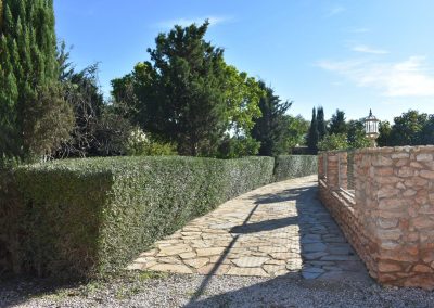 Camino con adoquines con seto a la izquierda y muro de piedra y valla a la derecha