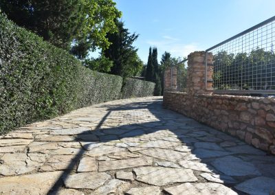 Camino con adoquines con seto a la izquierda y muro de piedra y valla a la derecha